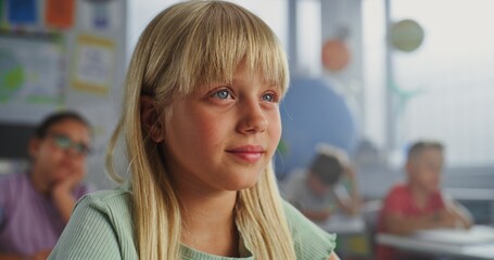 Close Up of Elementary School Girl Sitting at Desk, Raising Hand and Answering Teacher's Question...