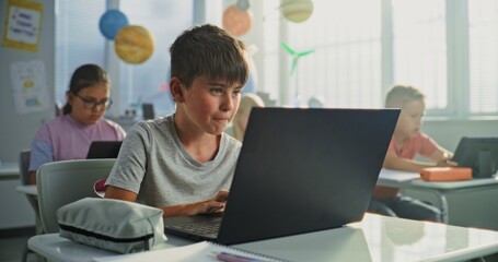 Computer Science Lesson: Primary School Boy Sitting at Desk, Using Laptop in Classroom. Group of...