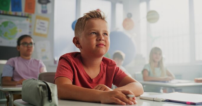 Elementary School Boy Sitting at Desk, Raising Hand and Answering a Teacher's Question During Geography Lesson. Team of Smart Diverse Kids Studying Environmental Science in Modern Colorful Classroom. - Powered by Adobe