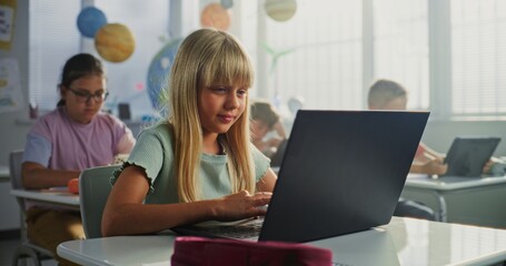 Elementary School Girl Sitting at Desk, Using Laptop in Classroom. Team of Smart Diverse Kids...
