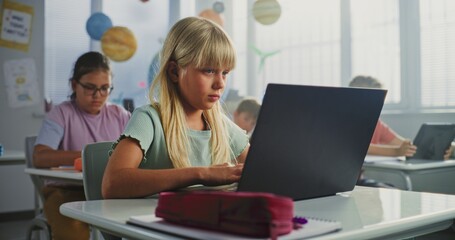 Computer Science Lesson: Primary School Girl Sitting at Desk, Using Laptop in Classroom. Group of...