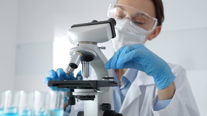 Woman science researcher wearing protective gear is checking microscope, analyzing blue test tubes...