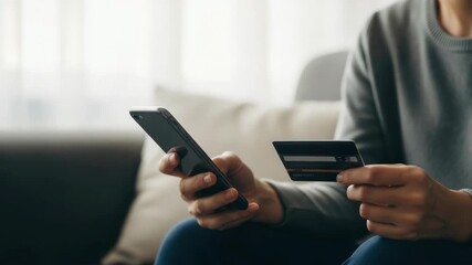 A person sitting on a couch using a smartphone to make an online purchase with a credit card while natural light filters through a nearby window creating a cozy atmosphere - Powered by Adobe