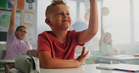 Elementary School Boy Sitting at Desk, Raising Hand and Answering a Teacher's Question During...