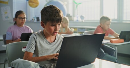 Elementary School Boy Sitting at Desk, Using Laptop in Classroom. Team of Smart Diverse Kids...