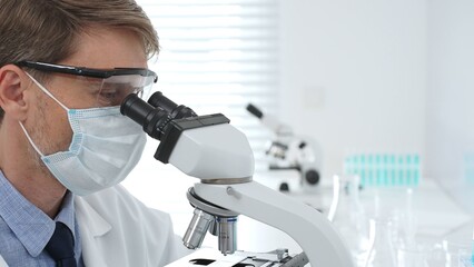 Scientist wearing mask and gloves using microscope doing scientific research in a laboratory with...