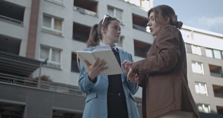 Two young businesswomen stand together, reviewing notes and having a discussion. One holds a notebook and the other gestures, potentially in a park or common area near an office building.