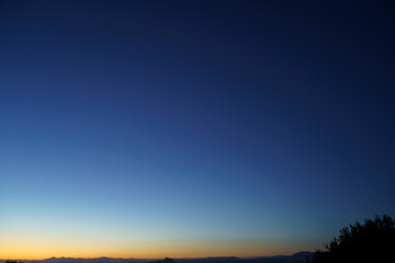 Silhouetted treetops and distant mountains lie under a vast, clear sky transitioning from the deep blue of twilight to the warm orange glow of dawn on the horizon.