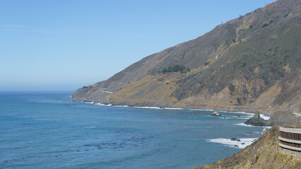 A high-angle, sunlit view of the rugged Big Sur coastline, showing the iconic Pacific Coast Highway as it winds along the cliffs above the turquoise waters of the Pacific Ocean.