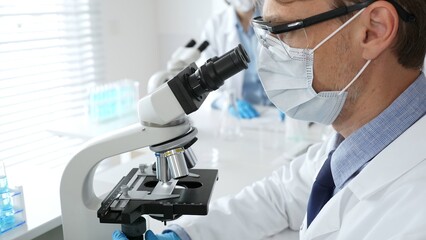 Masked scientist analyzing sample under microscope, colleague researching nearby in sterile medical...