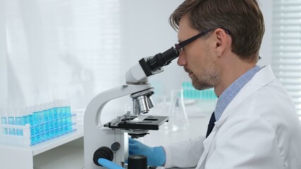Scientist wearing mask and gloves using microscope doing scientific research in a laboratory with...