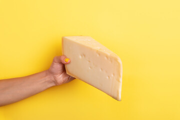 Close-up of a hand with yellow nails holding a large wedge of cheese against a vibrant yellow background, perfect for culinary and dairy themes.