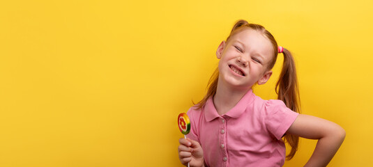 Cheerful young girl in pink shirt holding a colorful lollipop and smiling with eyes closed against yellow background.