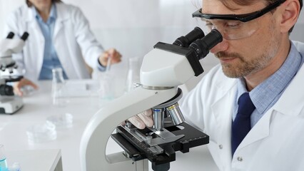 Medical scientist wearing gloves and protective glasses examining samples under a microscope in a...