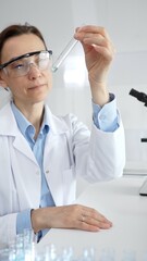 Female scientist wearing lab coat and protective glasses holding a test tube with blue liquid,...