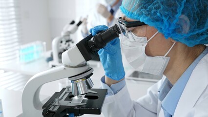 Female scientist wearing a mask and blue safety gear, using a microscope for research while a male...