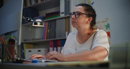 Elementary School Teacher Working on Computer, Checking Homework and Online Assignments During Distance Learning, Preparing Lesson Plan for Students. Woman Working at PC in Empty Classroom in Evening.