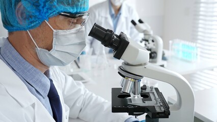 Senior male scientist wearing a protective mask, cap, and gloves, examining samples under a...