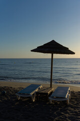 Two empty sun loungers and a straw umbrella sit on a sandy beach, with a calm ocean and distant mountains under a clear sky.