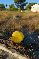 A weathered yellow buoy with a rusty chain rests on a piece of driftwood on a sandy beach, with tall grass in the background.