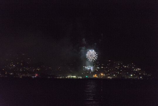 A series of vibrant fireworks light up the night sky over a body of water, with the sparkling lights of a city visible along the distant shore.
