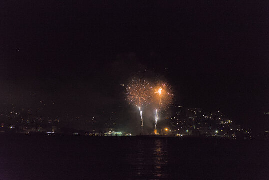 A series of vibrant fireworks light up the night sky over a body of water, with the sparkling lights of a city visible along the distant shore.
