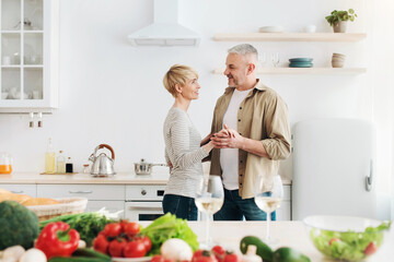 Romantic dinner in comfortable apartment, enjoy free time and healthy lifestyle. Cheerful senior man and lady dancing in light kitchen interior with bright colored vegetables on table, copy space