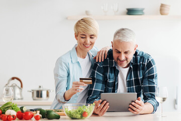 Positive emotions, shopping together, order online at home. Cheerful mature couple with credit card pay money through banking on laptop at kitchen interior, empty space