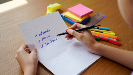 Child writing a checklist of school supplies on lined paper with pens, markers and sticky notes on a wooden desk. Back-to-school preparation