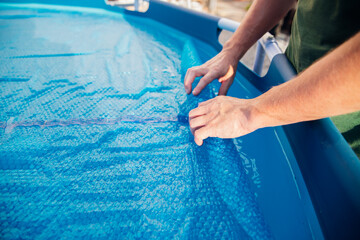 The blue pool cover is assembled into a roll by men's hands, the concept of summer recreation and...