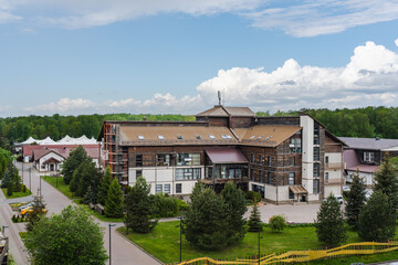 Modern multi-storey building with wood and white facade, surrounded by green trees and landscaped paths. Located in a peaceful area with forest in the background and a bright blue sky above.