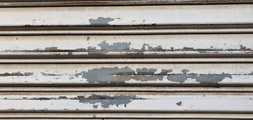 Close-up of an old metal rolling shutter with peeling white paint, scratches, rust stains, and weathered texture, showing urban decay and industrial wear