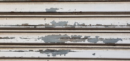 Close-up of an old metal rolling shutter with peeling white paint, scratches, rust stains, and weathered texture, showing urban decay and industrial wear