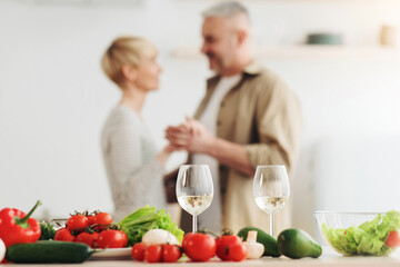 Romantic beautiful couple in modern cozy white kitchen room interior, celebrate holiday together at home. Glasses of wine on table with fresh vegetables, happy adult husband and wife dancing, blurred
