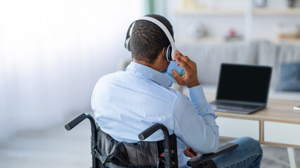 Back view of black guy in wheelchair wearing headphones, using laptop with mockup for design at home. New normal, be safe, remote job, online education, study and freelance concept