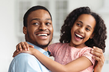 Beautiful black father and little daughter posing at home, embracing and laughing, looking aside....