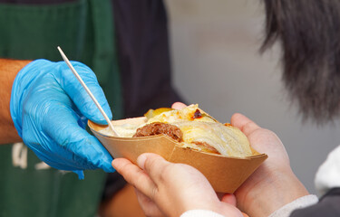 A gloved vendor serving customer melted raclette cheese over food at vibrant Prague farmers market. Authentic street food moment with rich texture and strong culinary appeal.