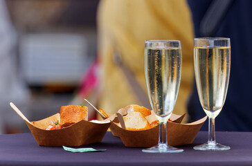 Two elegant flutes of sparkling wine stand beside bite-sized appetizers in paper trays at a sunny riverside farmers market on Prague Naplavka, evoking festive summer vibes.