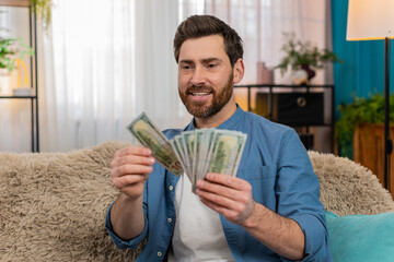 Young man at home holding a stack of dollar cash banknotes, counting money with excitement....