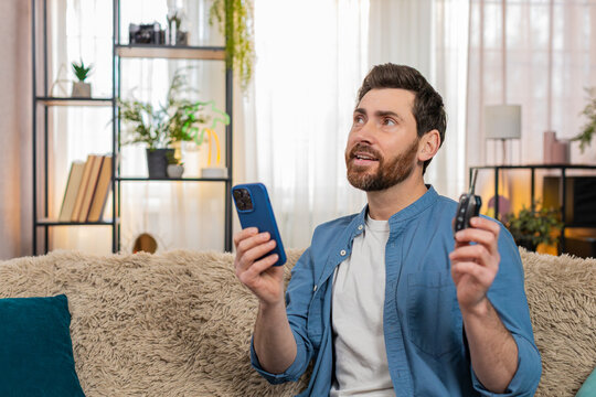 Young man at home intently checking a map on smartphone while planning a trip, gesturing in thought. Caucasian guy sitting on sofa, holding car keys, carefully considering travel route best directions