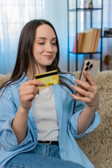 Young woman at home holding credit card and smartphone, happily making online purchase. Girl on...
