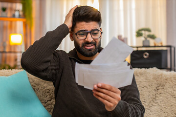 Indian man at home checks bills receipts with tense expression, nervously grabbing his head in...