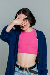 Young woman playfully peeks through hand formed like binoculars, smiling humorously, clearly indicating curiosity playful spying. Caucasian girl demonstrates humorous mood, isolated on gray background