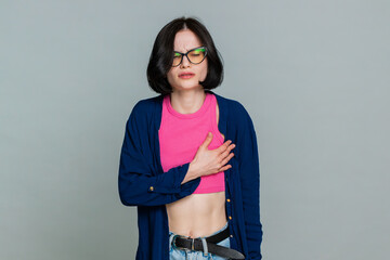 Young woman holds chest tightly with hands, looking downward sadly to clearly express emotional or heart pain. Girl demonstrates sorrowful feelings and deep emotional grief isolated on gray background