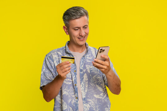 Middle-aged man holding smartphone and credit card while attentively entering payment information to make online purchase shopping. Young guy concentrated on transaction isolated on yellow background. - Powered by Adobe