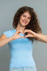 Young woman smiling warmly, making heart shape gesture with fingers expressing romantic love, care...