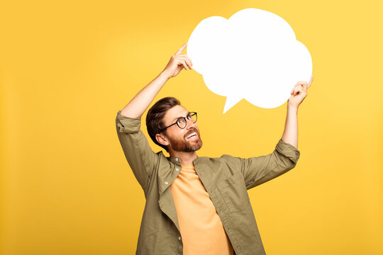 A middle aged man in casual attire stands against a vibrant yellow backdrop, smiling and holding a blank speech bubble above his head as if about to share a thought or message.