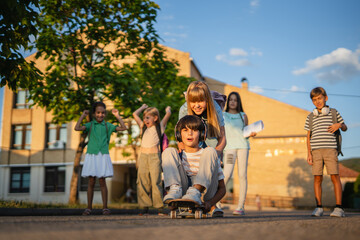 Schoolgirl pushing schoolboy sitting on skateboard after school