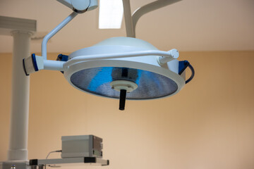 Modern surgical light fixture with reflective surface, suspended from ceiling in a clinical setting with beige walls and visible medical equipment.