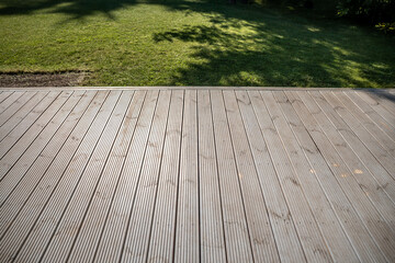 A wooden deck with visible grooves in the foreground, leading to a grass lawn. Tree shadows create...